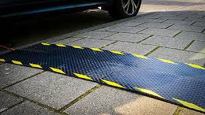 Een kabelmat ligt haaks over de stoep richting een geparkeerde auto. De kabelmat is zwart, heeft een antislipprofiel en is aan de lange zijden gemarkeerd met geel-zwarte lijnen.