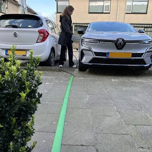 Een platte laadkabel ligt over de stoep richting een auto. De platte laadkabel is groen van kleur en heeft een antislipprofiel. 